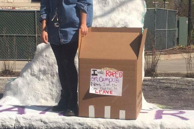 A female student stands by The Rock on Monday, displaying a cardboard box with a statement saying that she was raped. The student left campus on Tuesday.