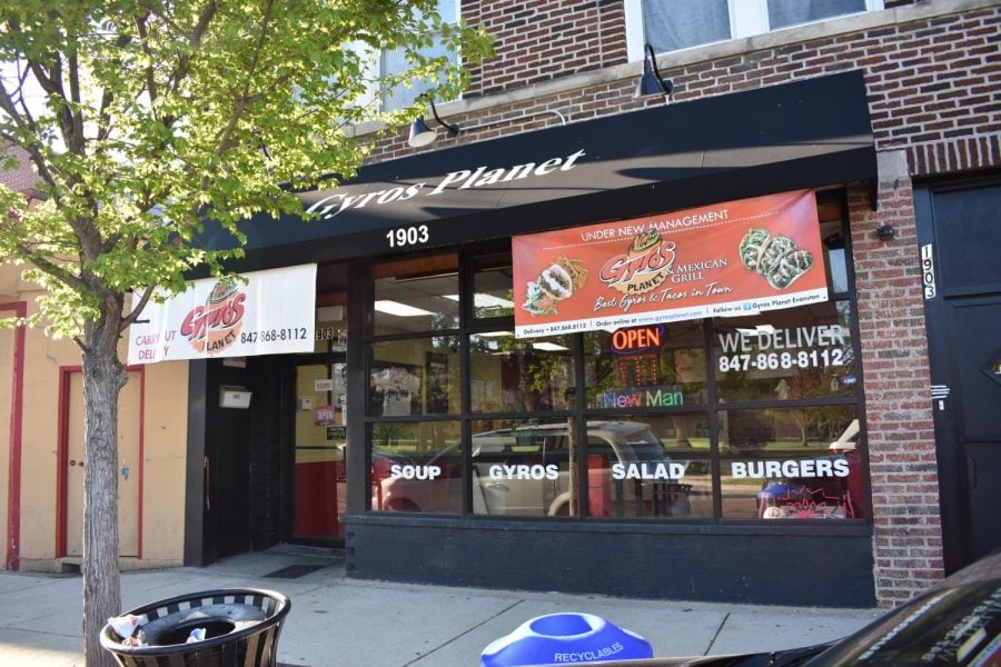 A storefront view of Gyros Planet, featuring signs detailing their specials and text on the windows reading, “Soup”, “Gyros”, “Salad”, and “Burgers”