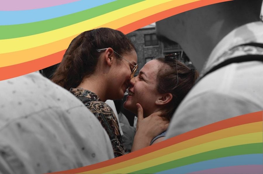 Two girls with wavy brown hair and sunglasses stand nose to nose. The shoulders and chins of other crowd members are visible in the foreground, with a rainbow across the top left corner.