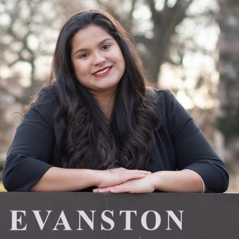 City clerk candidate Stephanie Mendoza stands smiling in a black blouse, with her arms resting on a brown sign that says “Evanston.”