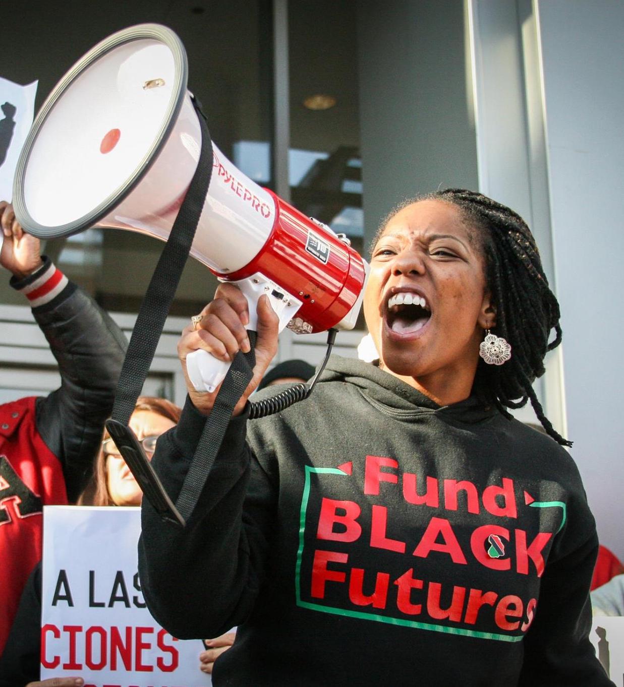 A+still+from+%E2%80%9CUnapologetic%E2%80%9D+of+Janae+at+a+protest%2C+speaking+into+a+megaphone.+She+is+wearing+a+t-shirt+that+reads+Fund+Black+Futures.