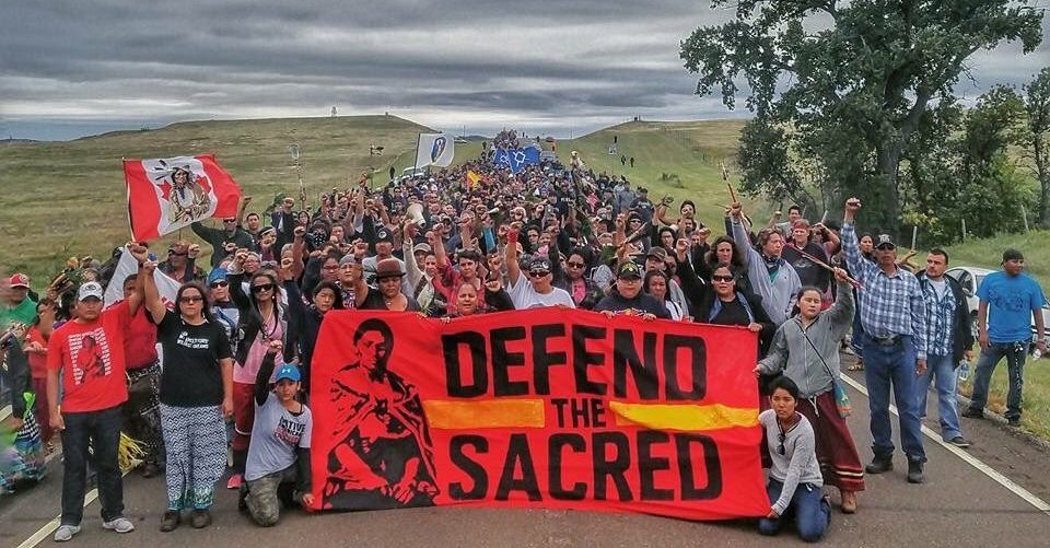 A+large+crowd+of+indigenous+people%2C+holding+a+red+sign+that+reads+Defend+the+Sacred.