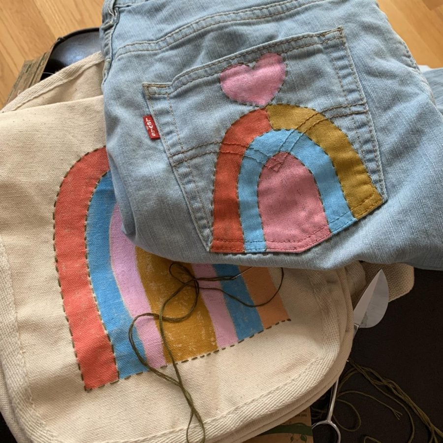A pair of folded blue jeans on top of a tan canvas bag, both with rainbows and hearts sewn on.