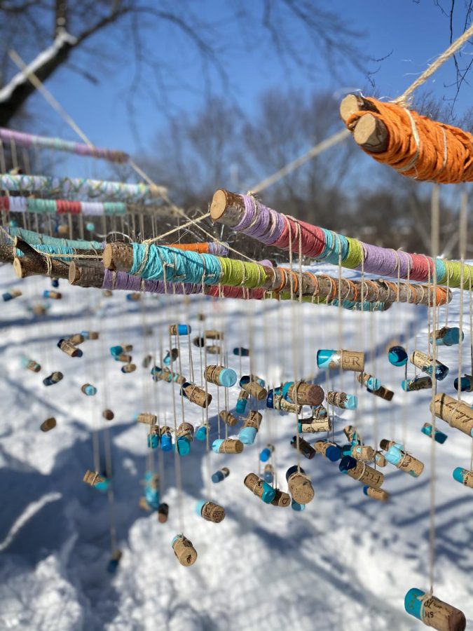 Sticks wrapped in colorful yarn are tied together with blue-dipped corks hanging beneath them on strings. There is snow in the background.