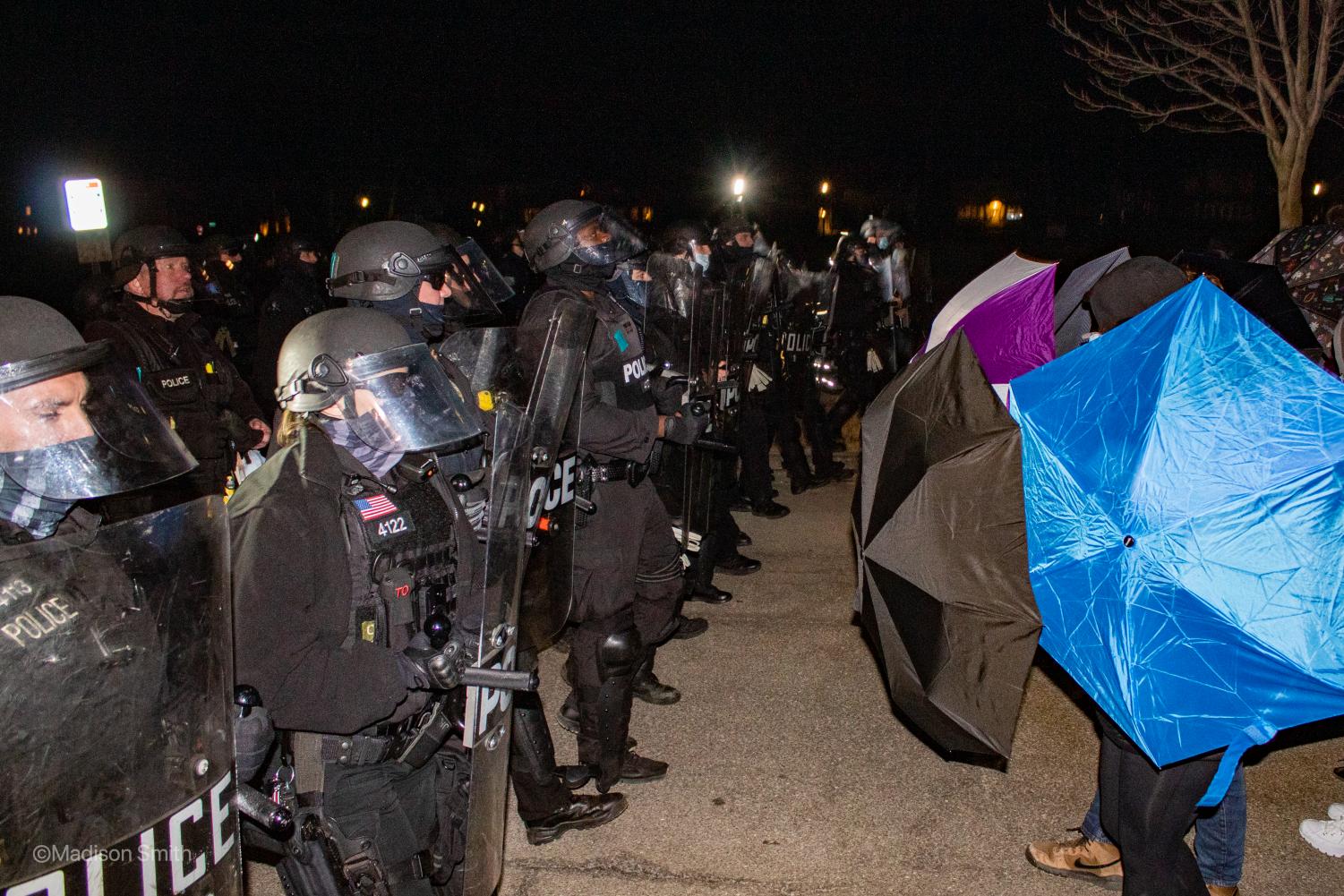 A+line+of+police%2C+fully+armed+in+riot+gear%2C+faces+off+with+protesters+who+are+protecting+their+faces+with+umbrellas.