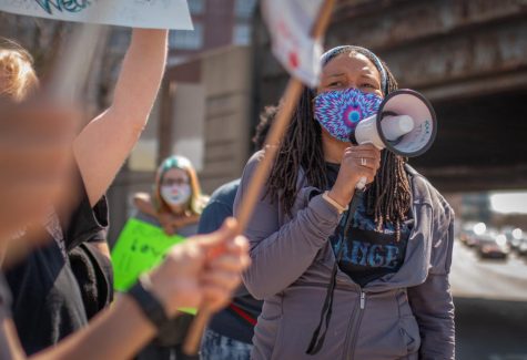 Karla Thomas, wearing a purple and blue tie-dye mask, speaks into a white megaphone while surrounded by participants carrying neon signs.