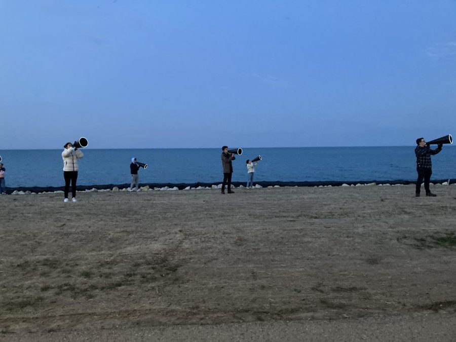 Several people stand several feet apart on the grass in front of Lake Michigan at sunset. They are holding large black megaphones with lights on the outer rim.