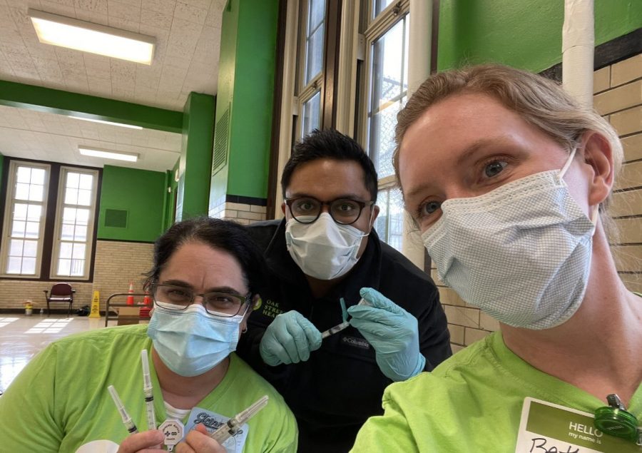 Three physicians at a vaccination site. The left one is wearing a green shirt and holding several vaccine needles, the middle one is wearing a black shirt and is also holding a needle. The right one is wearing a green shirt and taking the picture.