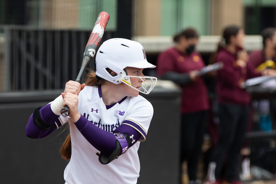 A softball player wearing white raises her bat, ready to hit the ball.