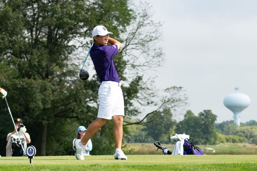 Golfer James Imai, wearing a purple shirt and white shorts, watches his shot while swinging through in front of a forest landscape and a distant water tower.