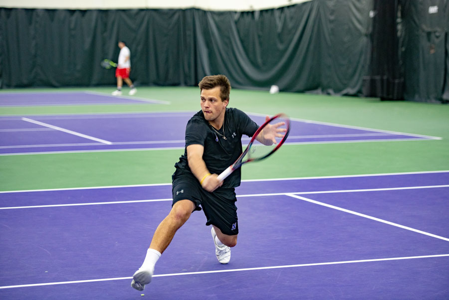 Northwestern men’s tennis player wearing all-black athleisure and holding a red and black racket returns a backhand shot on the Wildcats’ purple-and-white courts.
