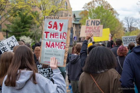 A protester, part of a group of protesters marching down Sheridan Road, holds up a sign with “Say their names” written in red text against gray paper.