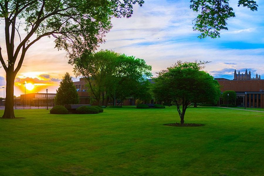 An open green field with trees in front of a school building. The sun is setting.