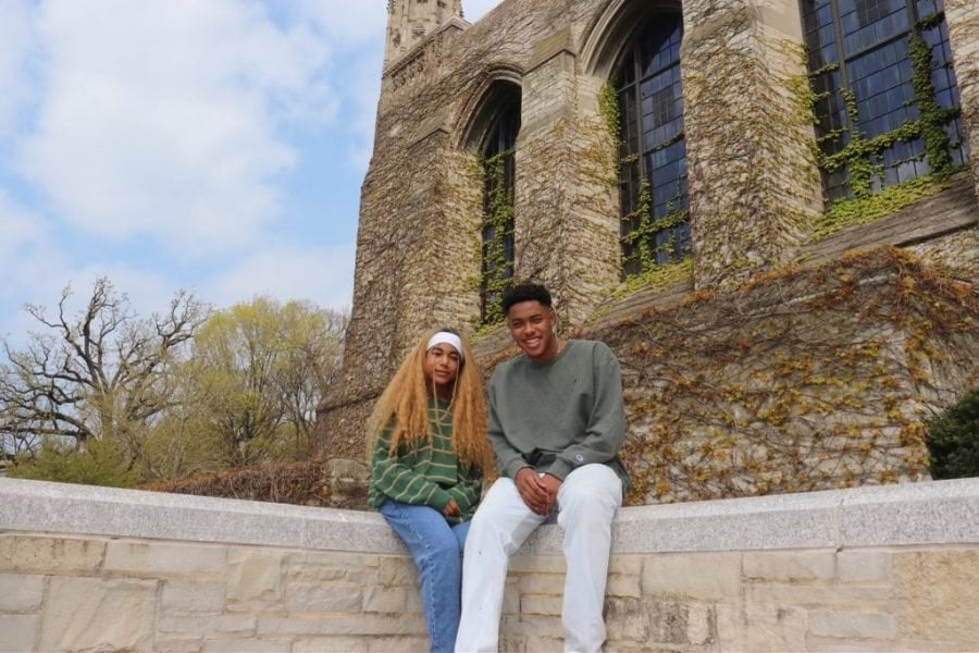 Christian Wade and Ada Ogbonna sit side by side on a concrete ledge in front of Deering Library. Ogbonna is wearing a long sleeve, striped green sweater with blue jeans and Wade is wearing white jeans with an olive green long sleeve shirt. There are trees behind them and a blue, cloudy sky and vines all across the library background.