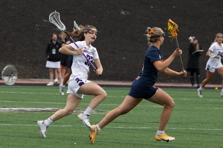 A+lacrosse+player+wearing+white+holds+her+stick+and+runs+down+field+to+guard+a+player+dressed+in+blue+with+an+orange+lacrosse+stick.