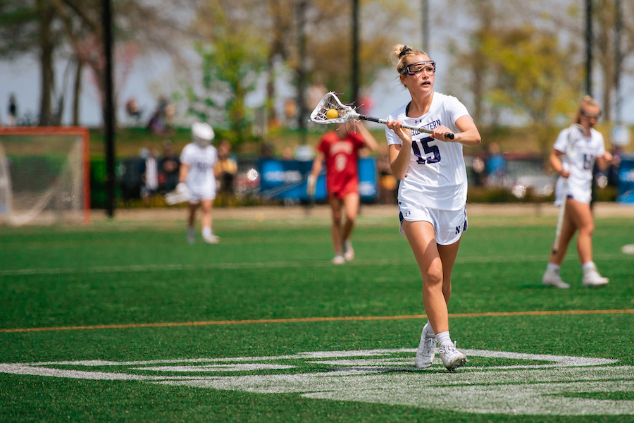 A lacrosse player wearing white with hair in bun runs with ball in her stick and looks to pass.
