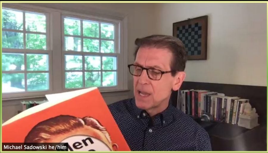 A+man+with+glasses+and+a+navy+collared+shirt+reads+from+a+book.+Part+of+the+book%E2%80%99s+jacket+is+visible.+He+is+in+a+room+with+a+window+and+books+in+the+background.