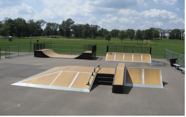 Brown skate ramps on grey concrete. A park with green grass and green trees is in the background.