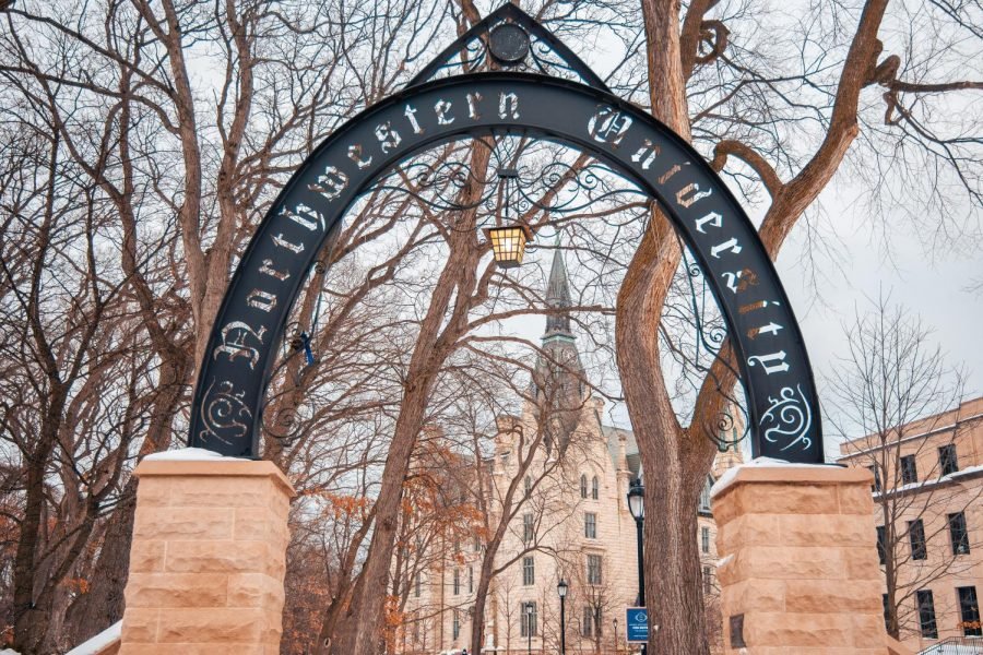 The Weber Arch. The dark gray arch is supported by two tan brick foundations. Behind the arch is the sky in light blue and brown, bare trees. Also in the background are campus buildings.
