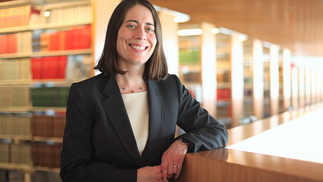 Hari Osofsky, in brown hair, is shown with her left arm perching. She is wearing a dark gray blazer and ivory shirt. In the background are slacks of books.