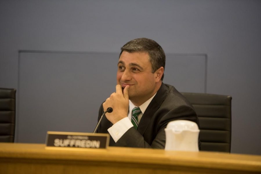 Ald. Tom Suffredin sits at the dais, wearing a grey suit with a green striped tie.