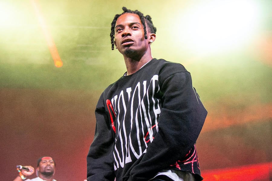 Playboi Carti wearing a black sweatshirt with white text on it while staring out from a stage.