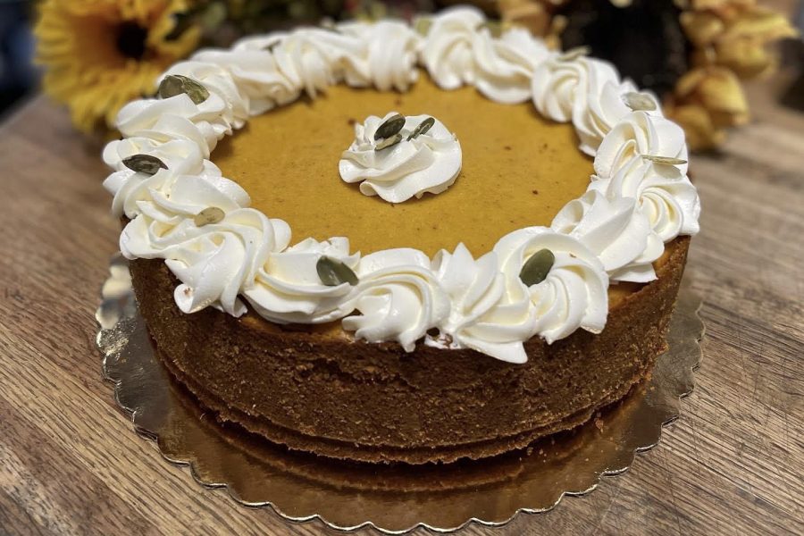 A brown cake with white frosting decorated with green seeds. There are sunflowers in the background, on a brown wood table.