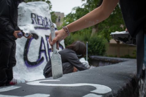 A person’s hand with a paint brush, stroking white paint on a short black wall. The wall encloses the Rock, which is painted white reads “Reform CAPS” and other event organizers painting the area.