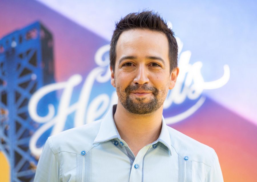 Lin-Manuel Miranda looks directly at the camera in front of an “In The Heights” poster.