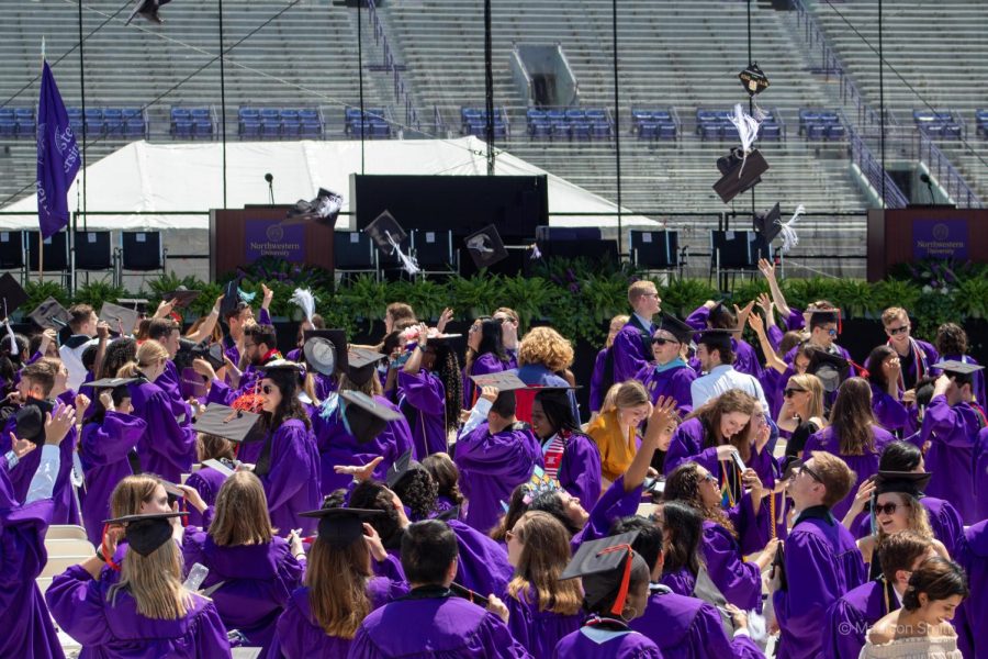 Crowd of people in purple graduation gowns gathered on a field, some throwing their caps into the air, with bleachers and a podium in the background.