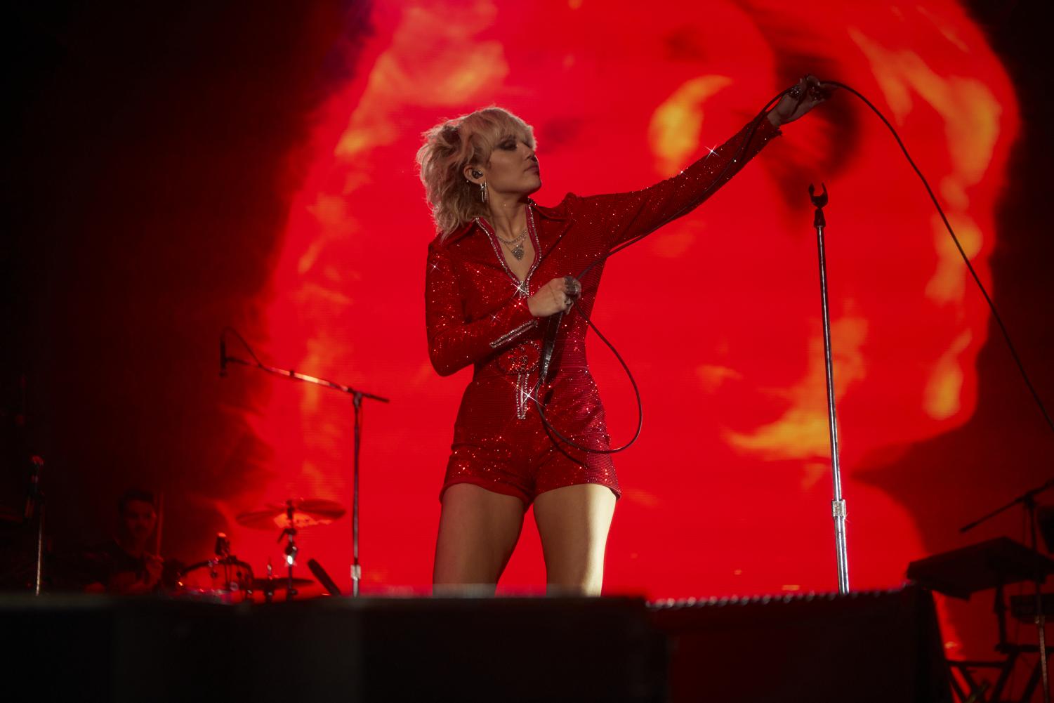 Miley+Cyrus%2C+wearing+a+red+sequined+jumpsuit%2C+looks+out+into+the+crowd%2C+arm+extended%2C+while+holding+her+mic.