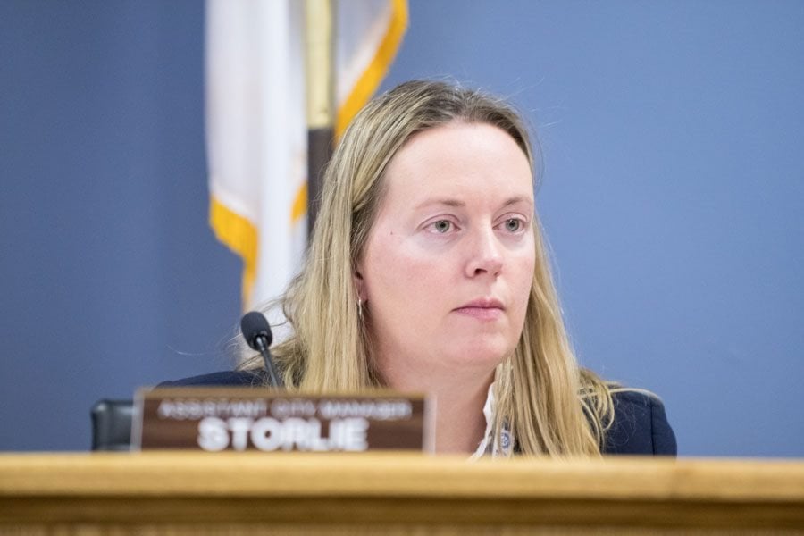A woman with blond hair sits behind a microphone and a brown name tag that reads “Storlie.”