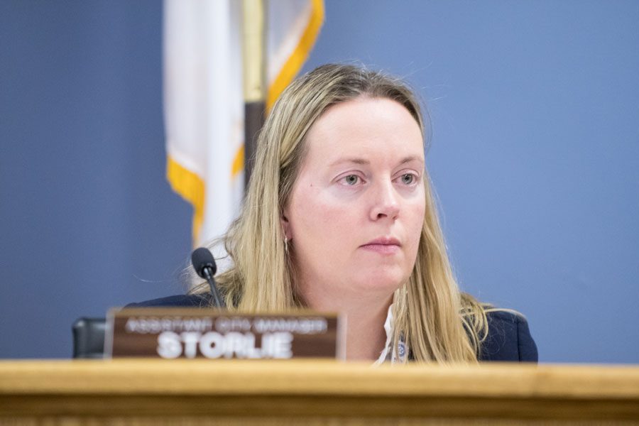 Erika Storlie sits at the dais, a flag behind her, wearing a suit jacket and white shirt.