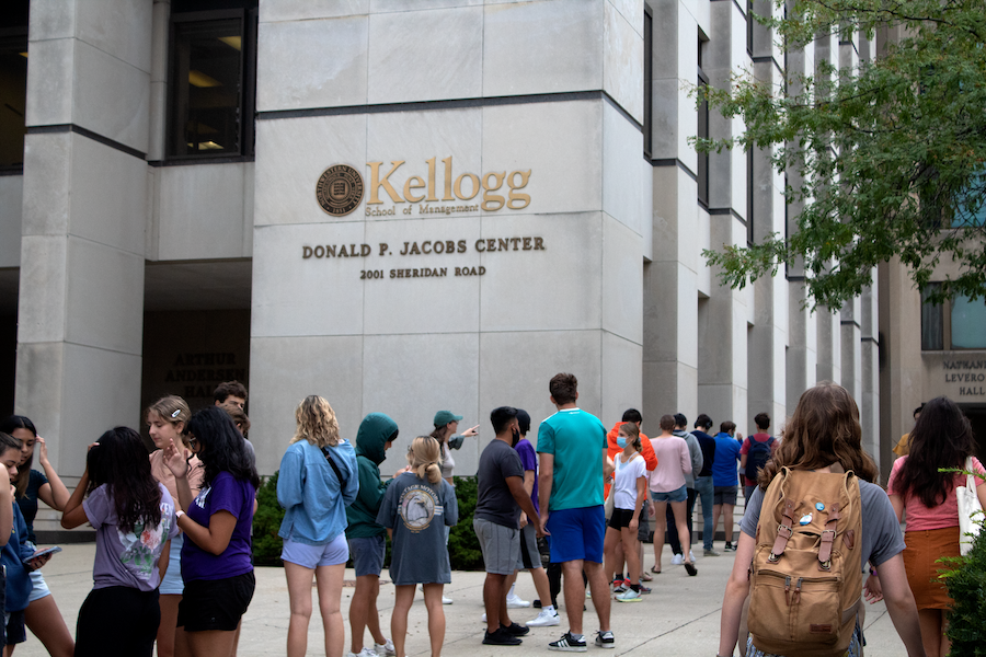 Kellogg School of Management in golden letters on a tan building with several students talking and standing in a line.