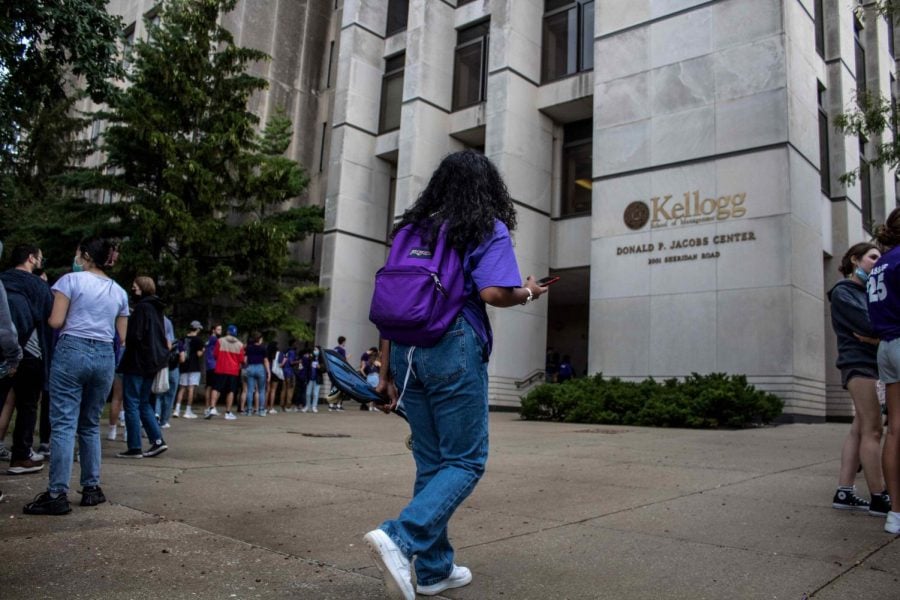 Person with purple backpack in front of the Jacobs Center is walking. A line of students trail the testing center, a white building with golden letters.