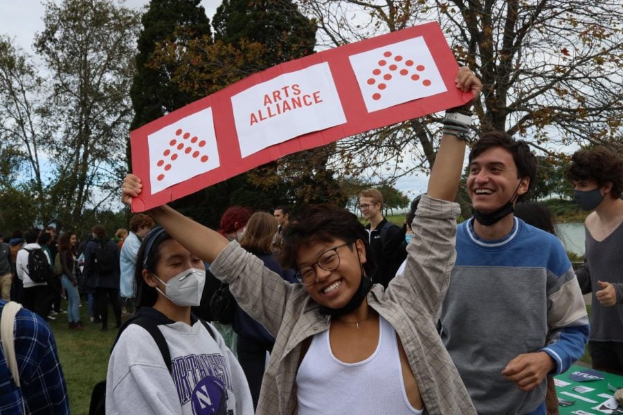 A student waves a red banner that reads “Arts Alliance” above his head, with two other students behind him.