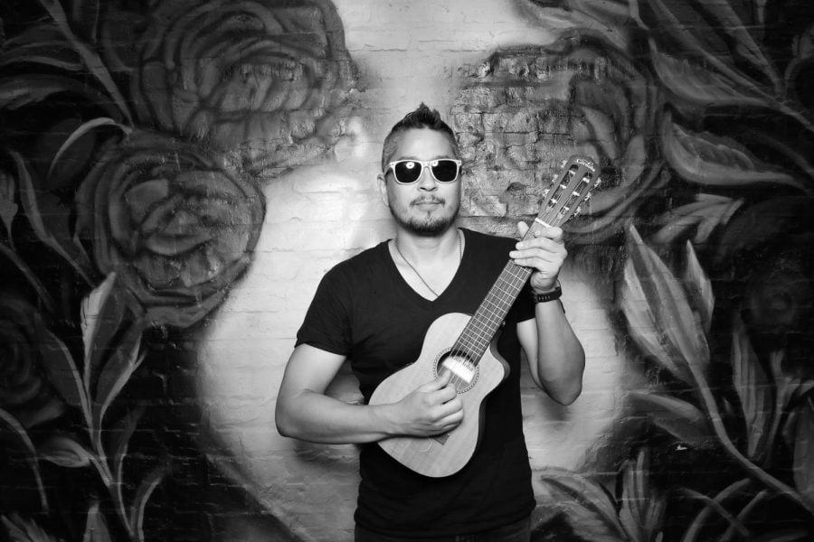 Local musician Cary Kanno dons a black t-shirt and sunglasses as he holds a small guitar in front of a black-and-white wall with flower designs.