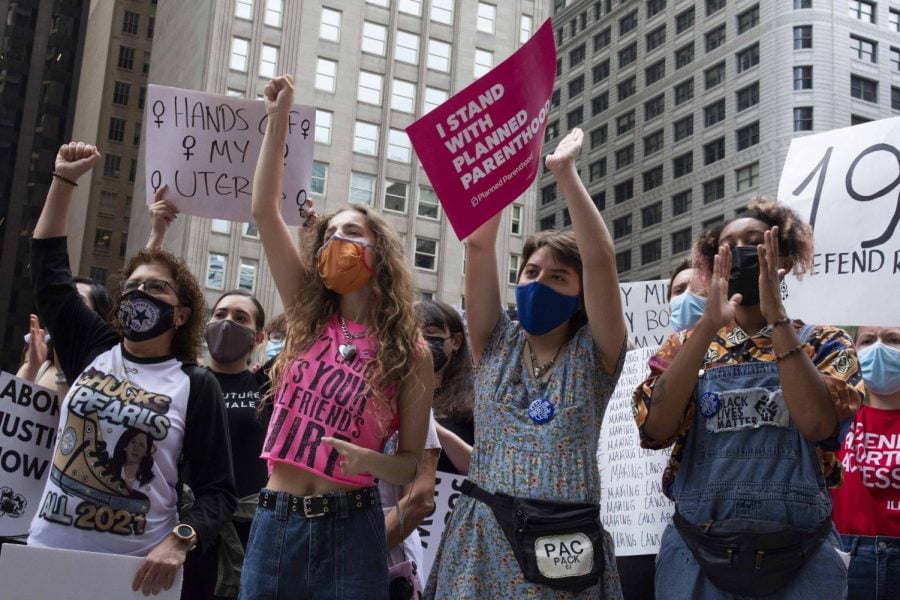A woman in a pink shirt stands next to another woman in a blue dress, both of whom are pumping up their fists among others in the crowd among them. The woman on the right holds up a pink sign saying “I stand with Planned Parenthood” in capital white letters.