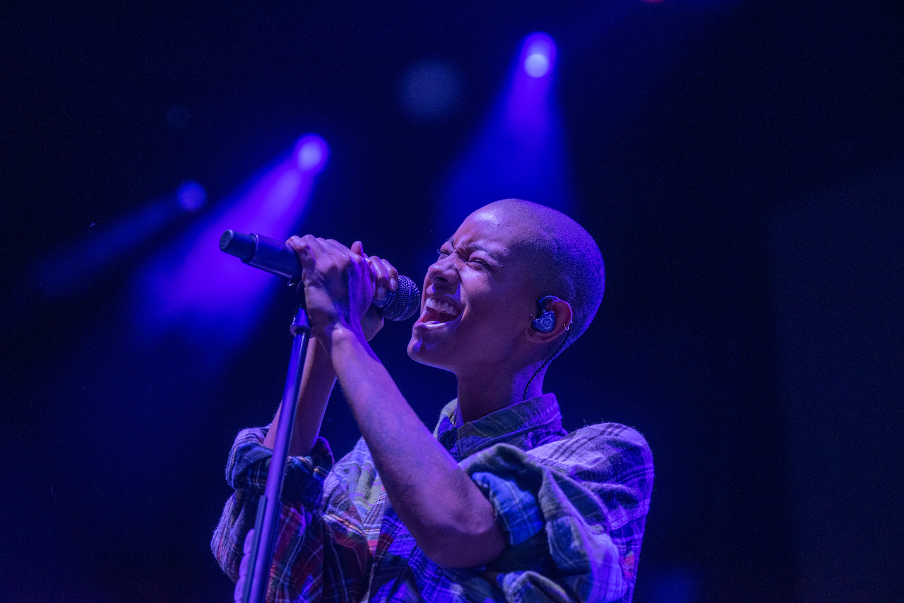WILLOW is illuminated in blue stage-lighting as she sings into a microphone.