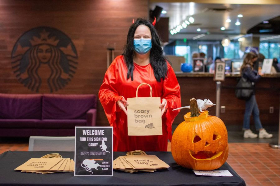 A person in a red devil costume holds a brown bag with the text “scary brown bag” on it behind a table with a pumpkin and more brown bags.