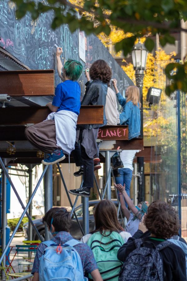 Four people sit up on diagonally crossed metal poles, drawing on the plywood the structure is supporting. Below them, other students watch.