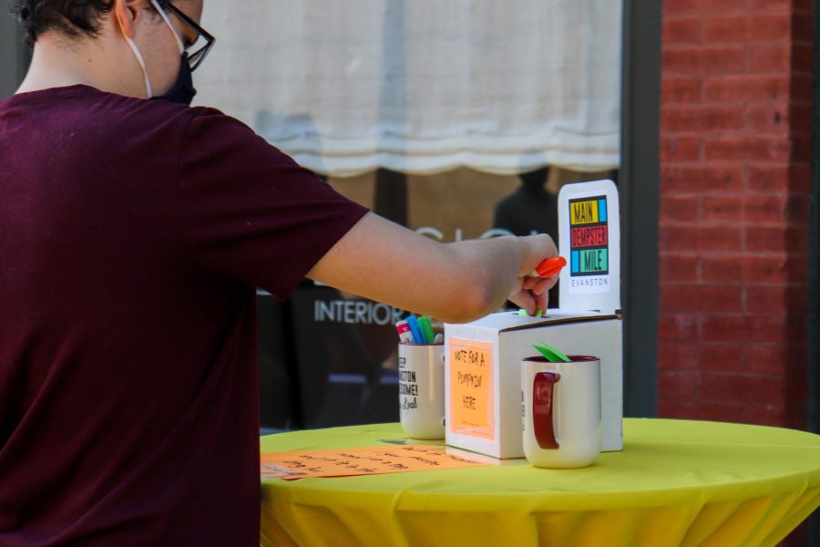 A visitor wearing a maroon shirt inserts their ballot into a white box with an orange label that says, “VOTE FOR A PUMPKIN HERE.” There is a red-and-white cup with colorful paper next to the ballot box so visitors can cast their votes and another cup on the other side of the box with color sharpies. There is an orange paper in front of the box that explains how to vote.
