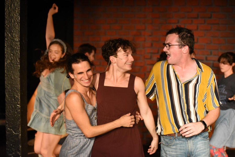Three people stand on stage, hugging and smiling. Behind them are more people dancing in front of a brick background.