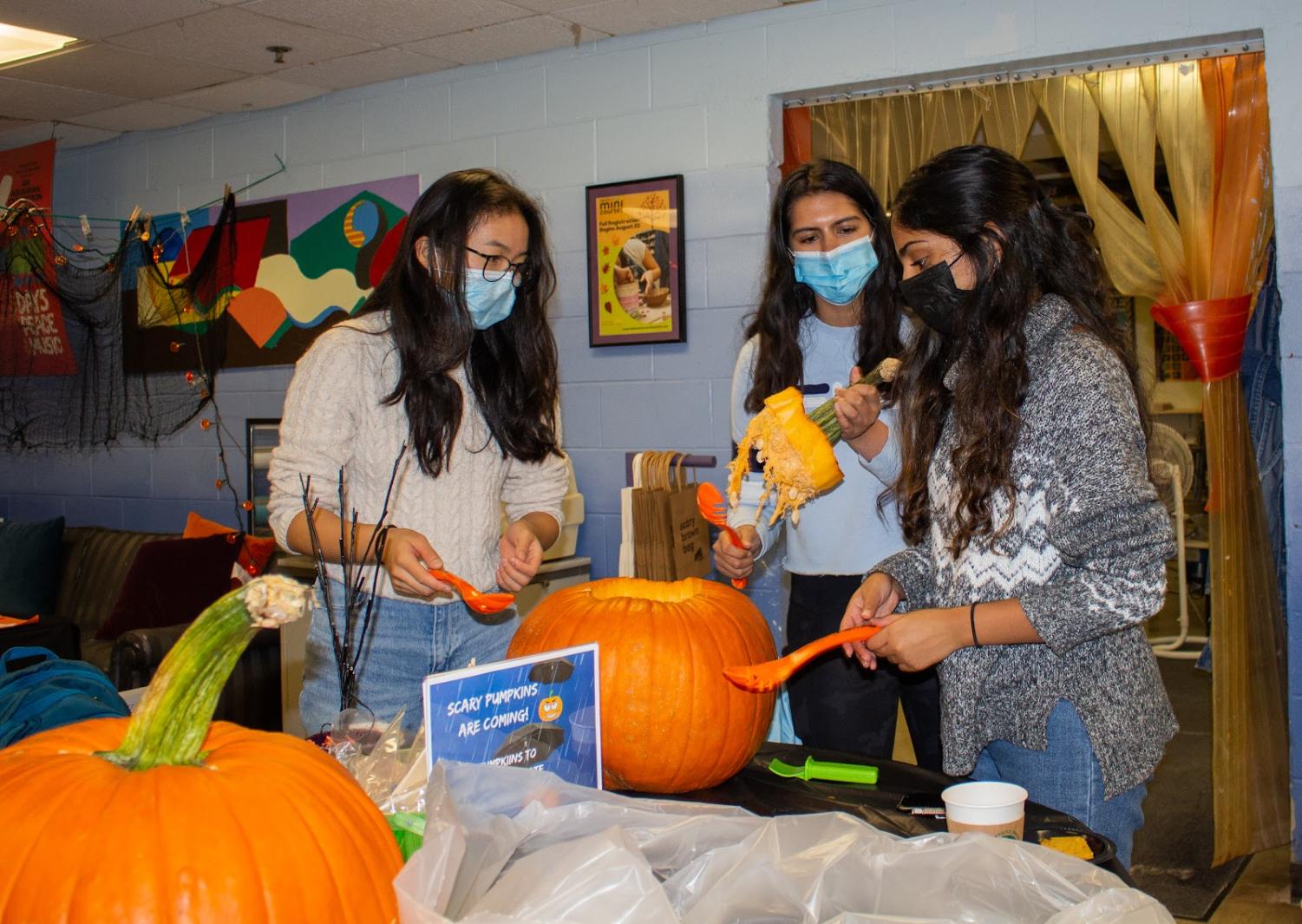 Three students holding pumpkin-carving tools around a pumpkin.