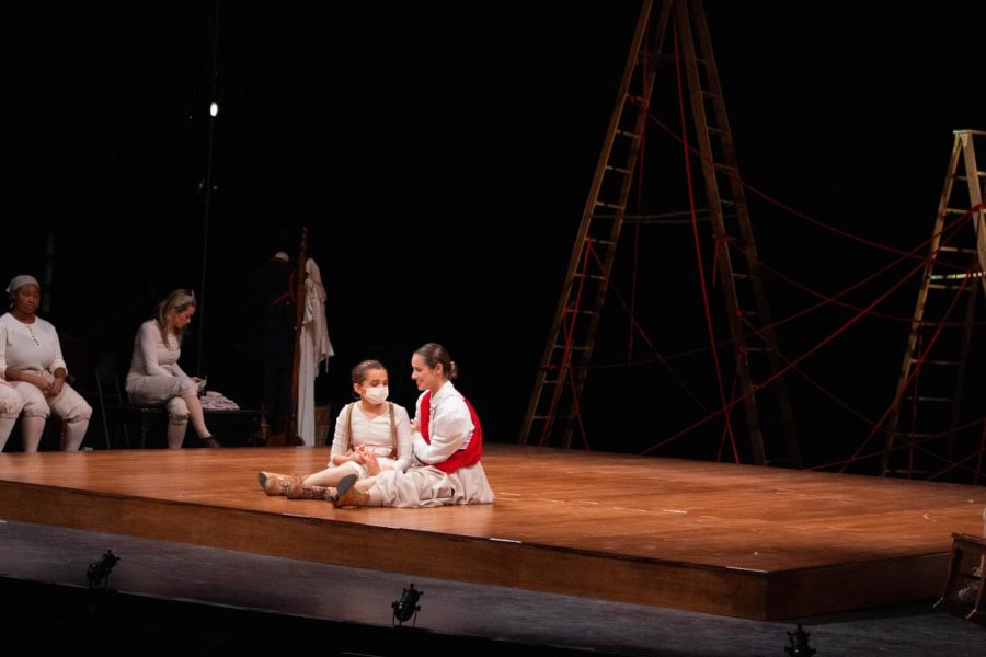Two characters sit together closely on the floor of the stage. They’re both in white-and-khaki clothing, and one is smiling at the other.