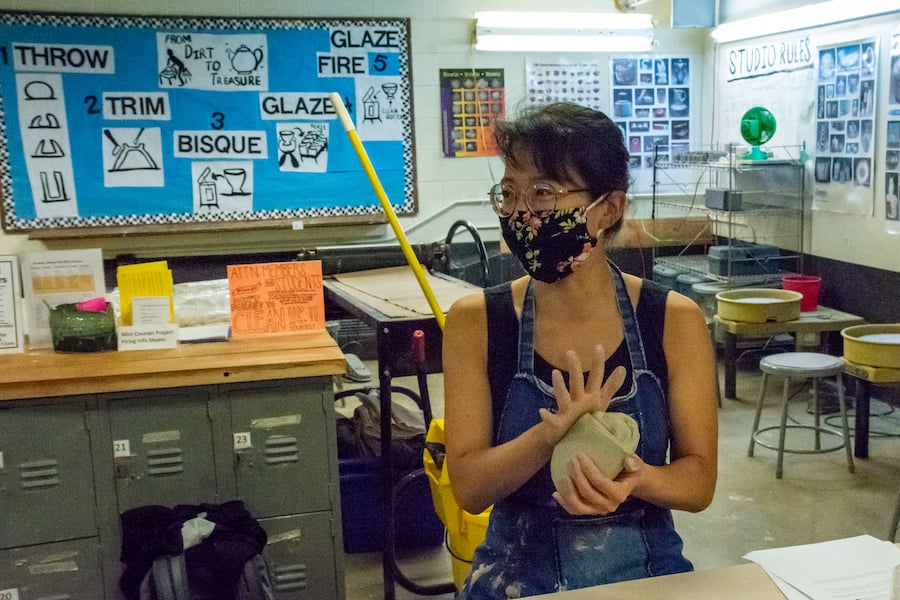 A woman wearing glasses, a black floral mask and denim overalls with a black tank top underneath kneads a gray ball of clay in an art classroom.
