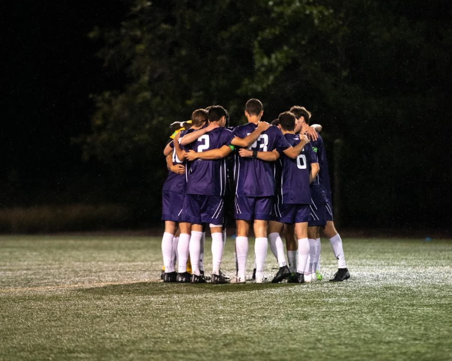 A group of players in purple jerseys, purple shorts and white socks huddle together on a green field.