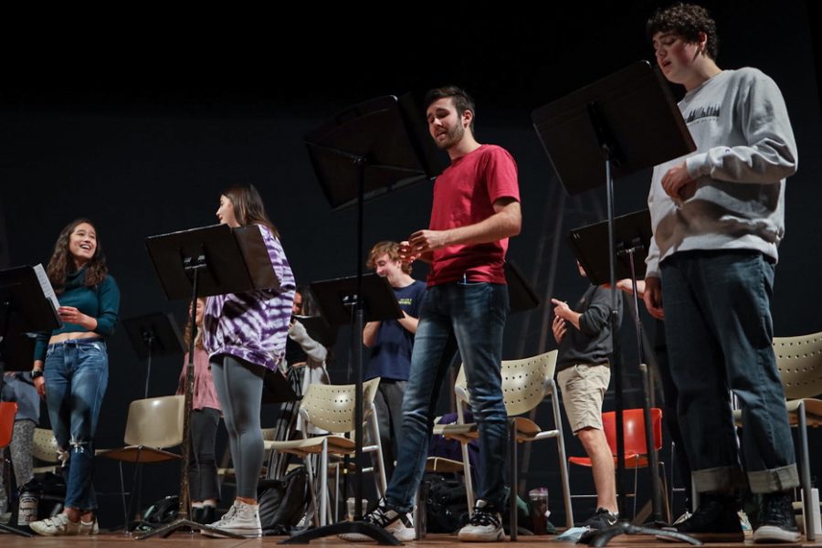 Students stand on an elevated stage in front of music stands and sing.