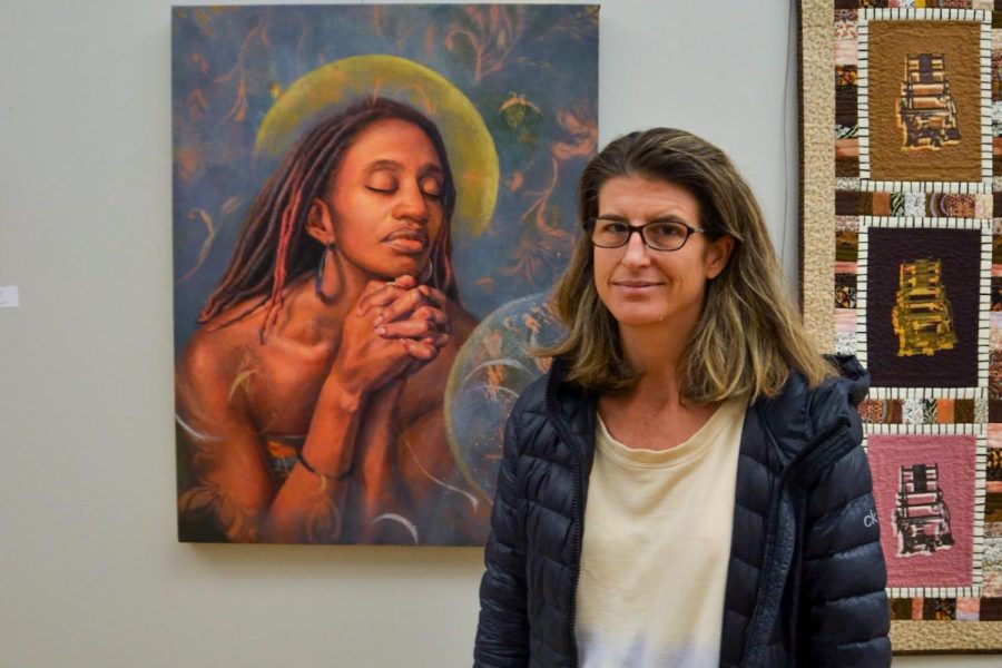 A woman with brown hair, glasses, a long black coat, and a white shirt that fades into blue, pink and green stands to the right of her painting of a Black woman in the middle of prayer.