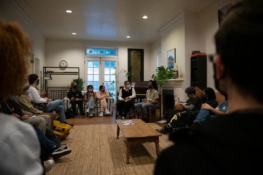 Two people sit in conversation in the living room of University Christian Ministry. They are surrounded by people sitting on couches and foldable chairs.